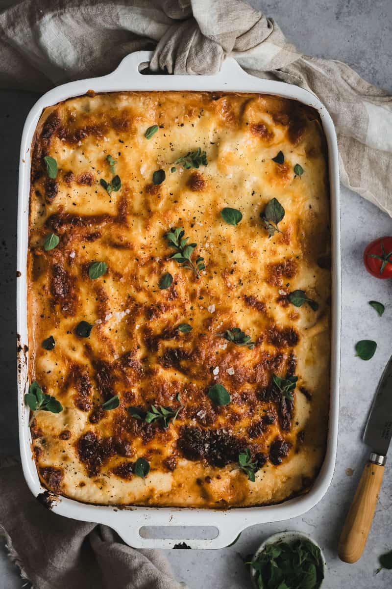 A whole golden, cheesy lasagna just out of the oven in a white roasting dish. It has been scattered with fresh herbs. Fresh baby tomatoes and a small knife sit beside.