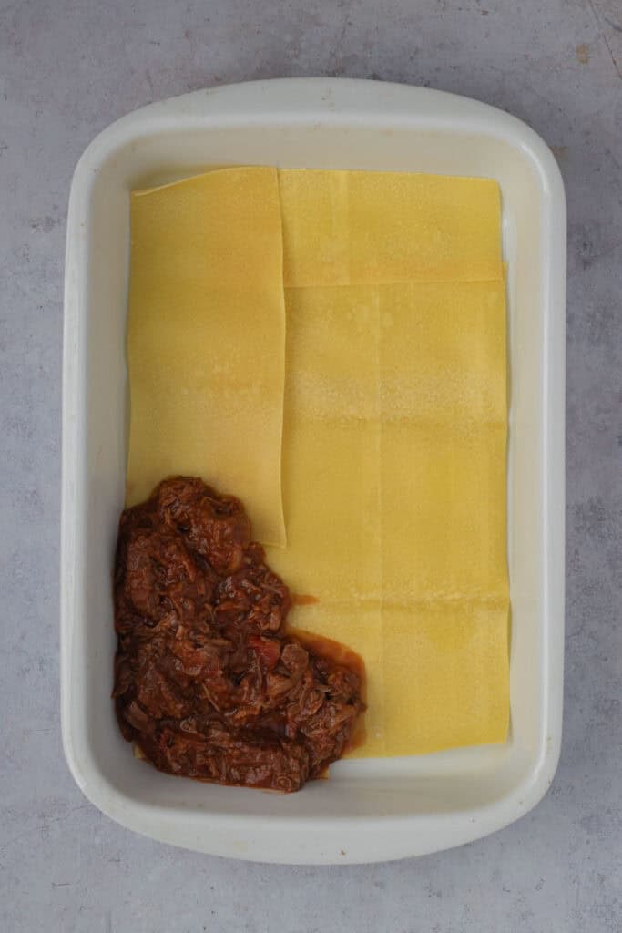 Lasagna pasta sheets line a white roasting pan. One corner has a little ragu on top of the pasta.
