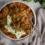 A bowl of slow cooked beef curry on a bench. A grey cloth sits beside along with some fresh coriander. A serving fork and spoon sit on the side of the bowl.