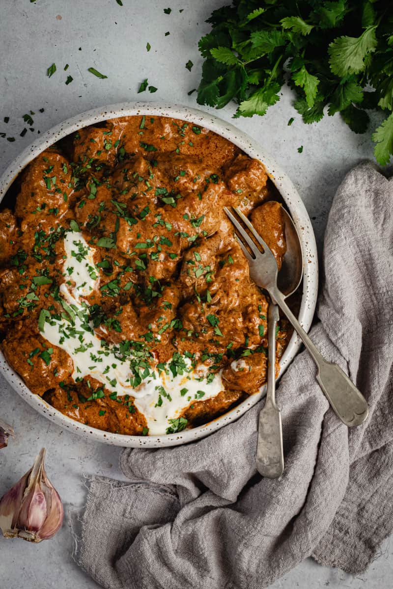 A bowl of slow cooked beef curry on a bench. A grey cloth sits beside along with some fresh coriander. A serving fork and spoon sit on the side of the bowl.
