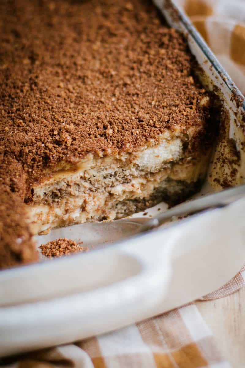 A closeup of a cast iron baking dish full of tiramisu with a slice eaten out of it.
