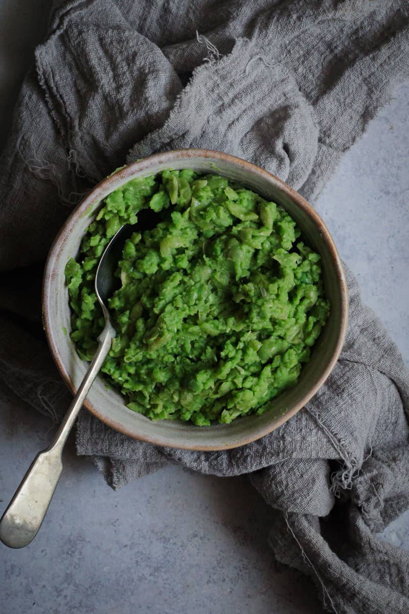 Close up of a bowl of mushy peas on a grey background. The bowl sits on a grey cloth.