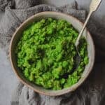 A bowl of bright green mush peas sit on a grey cloth. A spoon is in the bowl.