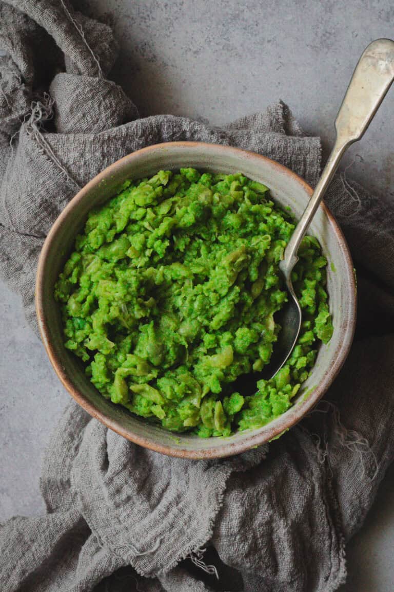 A bowl of bright green mush peas sit on a grey cloth. A spoon is in the bowl.