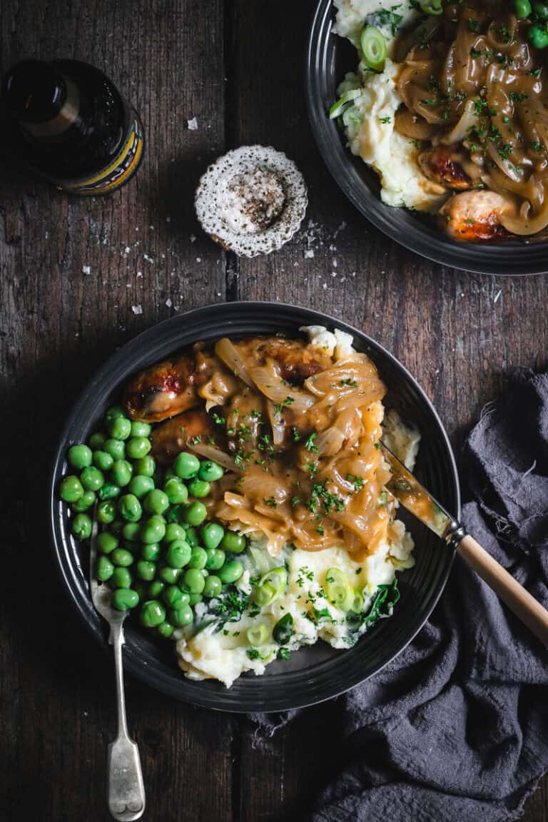 Two grey plates of colcannon, sausages peas and onion gravy sit on a timber table. A bottle of Irish cider and a pot of salt sit beside.