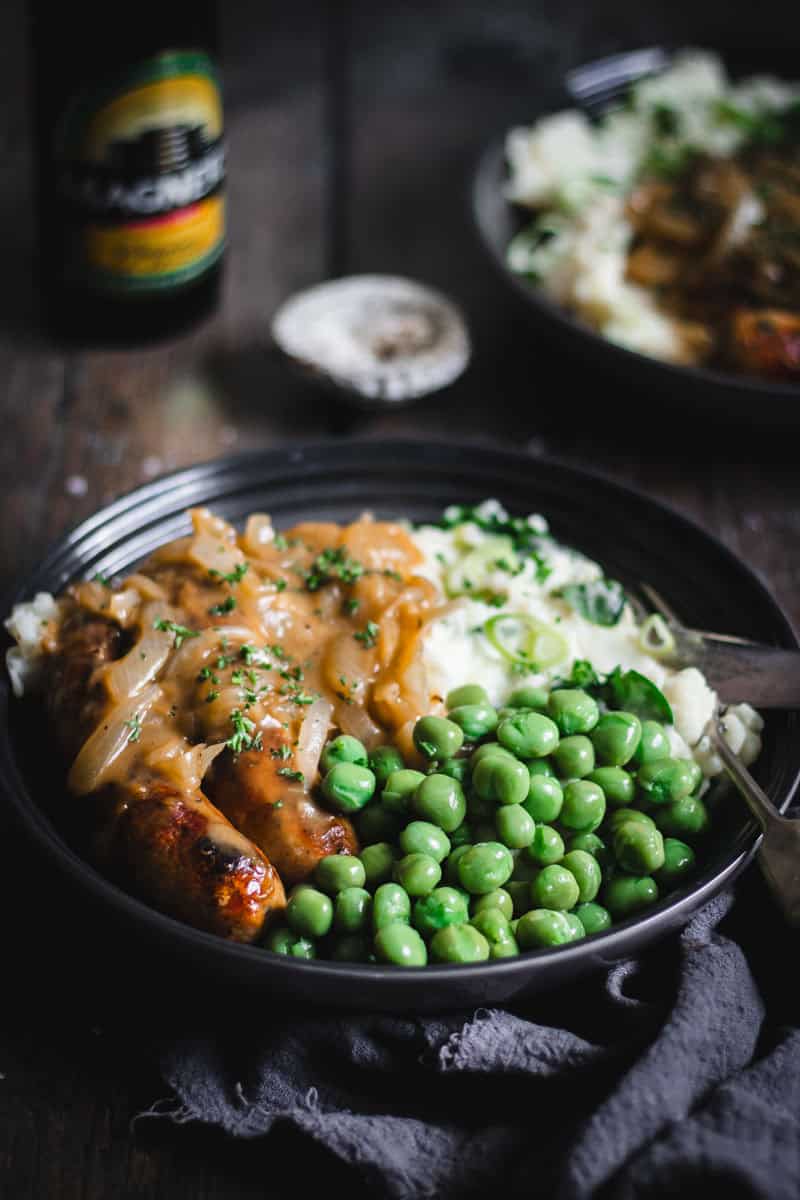 Two large shallow bowls of colcannon, sausages with onion gravy and marrowfat peas sit on a timber table.