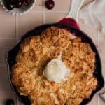 A white skillet with a baked cherry cobbler in it. A scoop of vanilla ice cream is melting on the top of the cobbler.