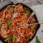 A black skillet of pork bolognese. A fork and a spoon have spaghetti twisted around them and there is a small bunch of basil on the bench.