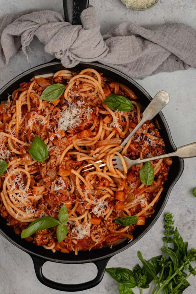 A black skillet of pork bolognese. A fork and a spoon have spaghetti twisted around them and there is a small bunch of basil on the bench.