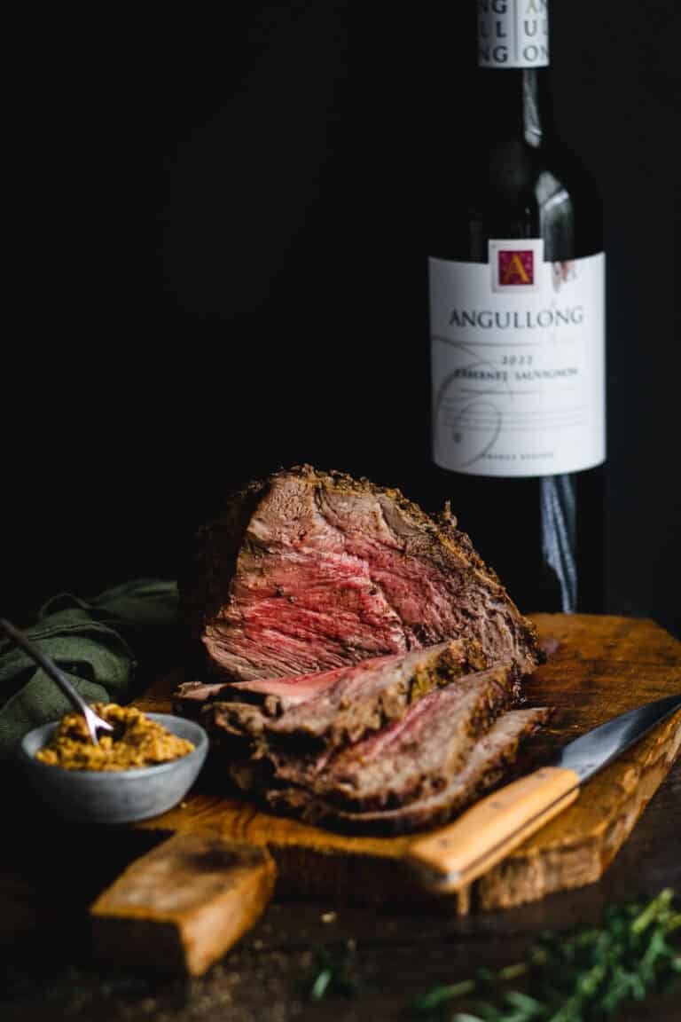 A rare medium rare roast beef being carved on a timber board. A bottle of Angullong red wine sits behind.