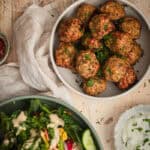 A bowl of meatballs sits on a timber table with a side salad a glass of white wine.
