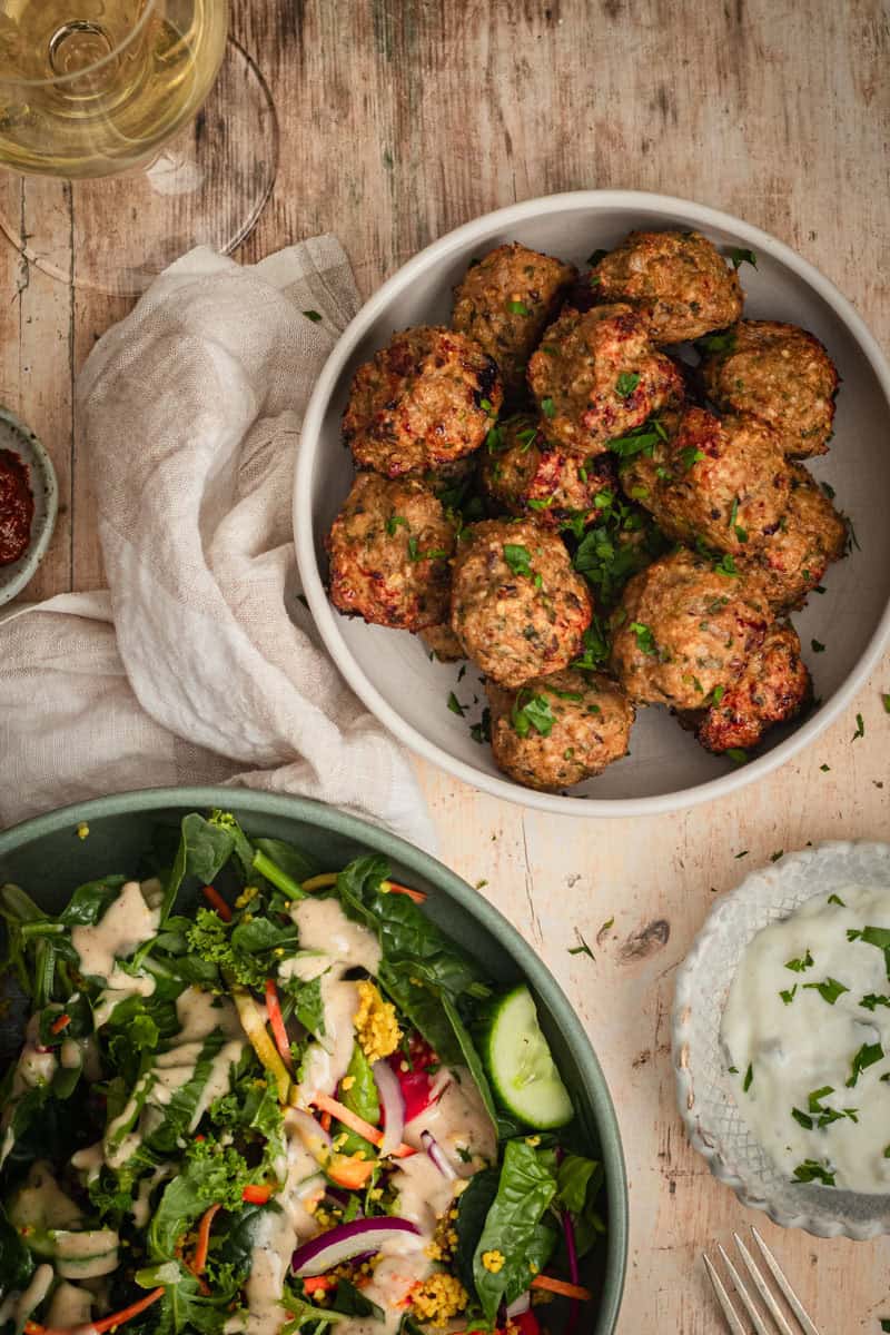 A bowl of meatballs sits on a timber table with a side salad a glass of white wine.
