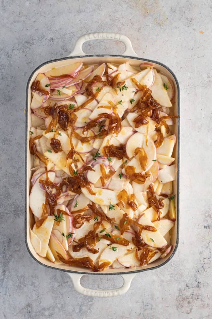 A cream cast iron roaster with a prepped potato bake in it just before it goes into the oven.