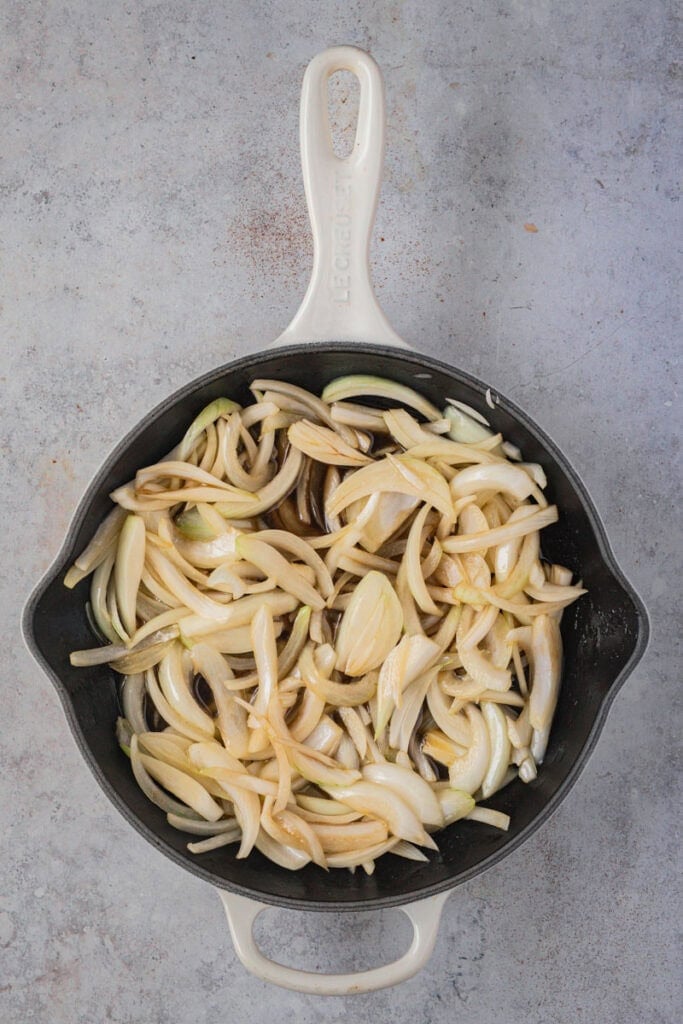 A cream colored cast iron skillet full of raw sliced onion, balsamic vinegar and brown sugar.
