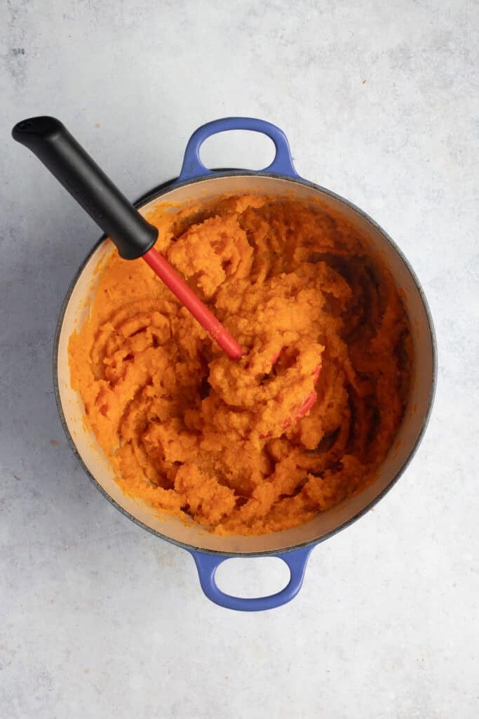 A red potato masher sits in a pot of mashed sweet potato.