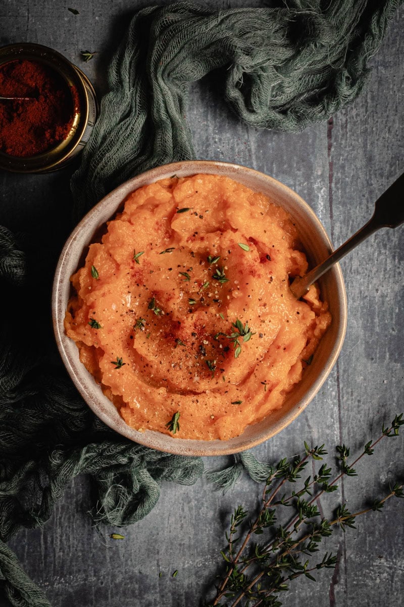A bowl of sweet potato mash with a spoon in it sits on a grey board along with some fresh thyme.