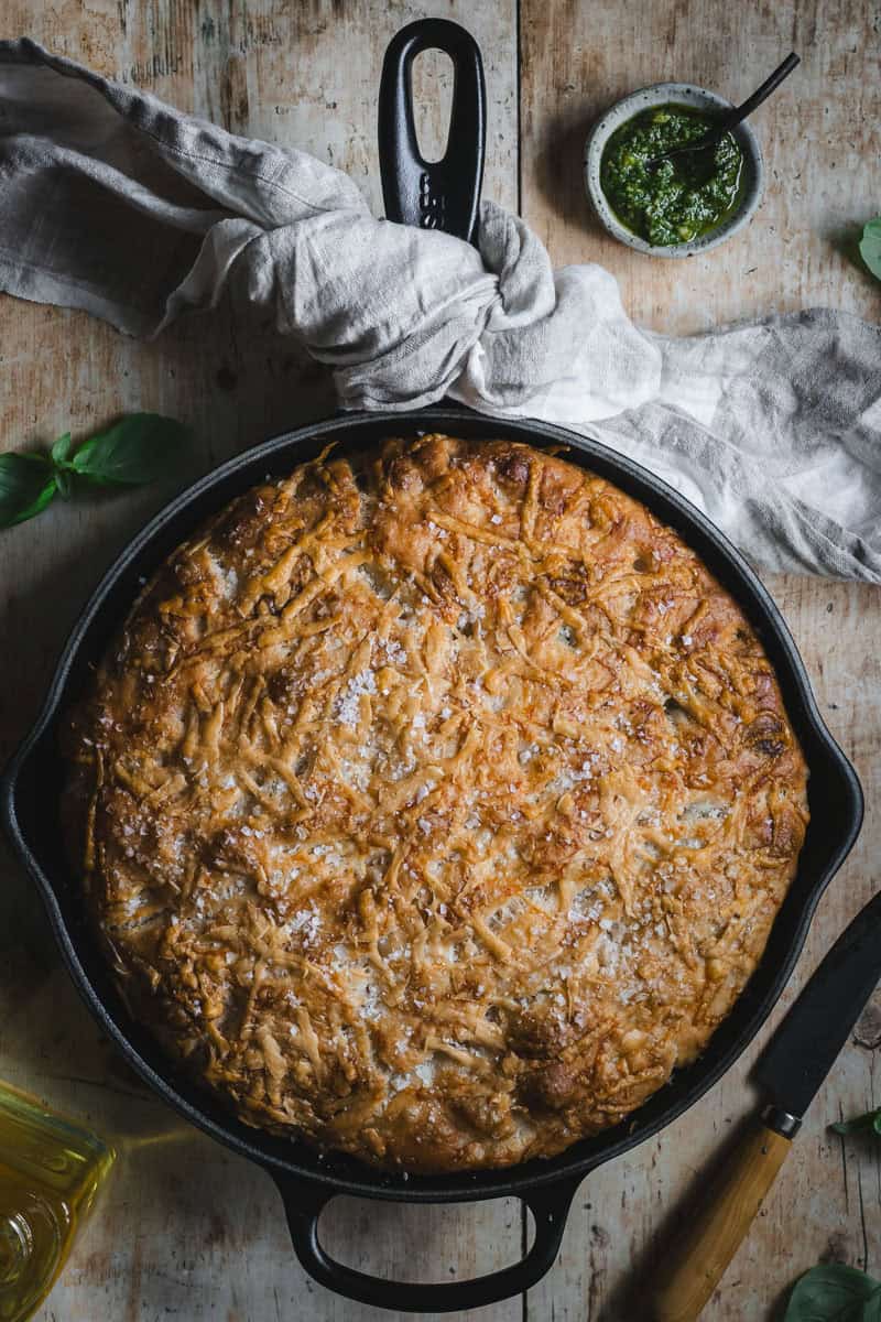 A black cast iron skillet with a pesto focaccia in it. A tea towel is tied around the handle.