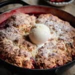 A strawberry cobbler in a shallow Dutch oven with a scoop of vanilla ice cream in the center.
