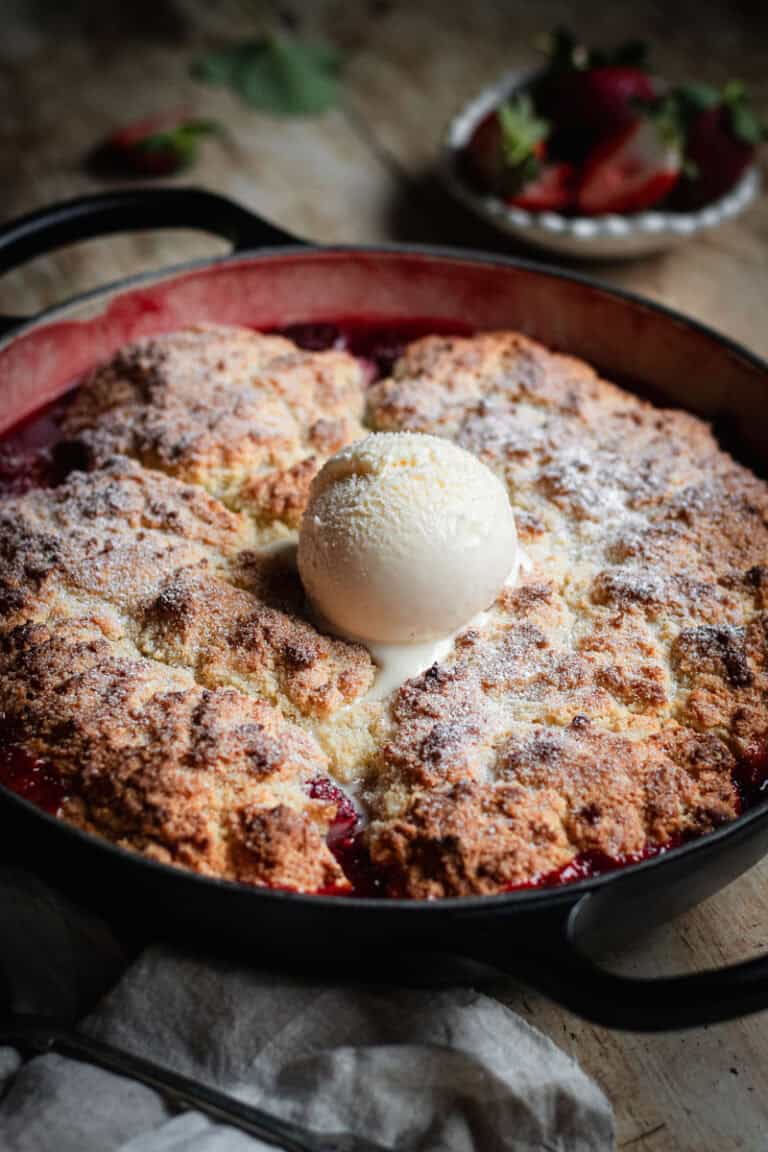 A strawberry cobbler in a shallow Dutch oven with a scoop of vanilla ice cream in the center.
